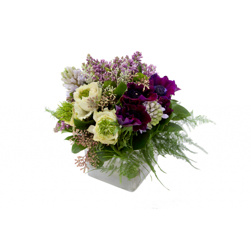 Flower arrangement in a low, square, white, ceramic vase, lilac, lavender hyacinths, purples anemones, white ranunculus, seeded eucalyptus and asparagus fern.
