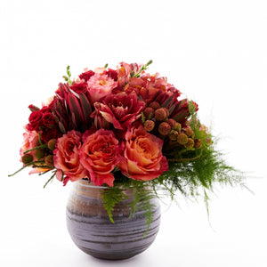 Flower arrangement in a low, round, brown, ceramic vase, coral roses, red spray roses, coral snapdragons, safari sunset and asparagus fern