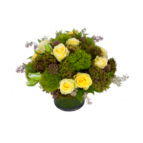 Flower arrangement in a low, round, clear, glass vase, yellow roses, green dianthus, green moss, scabiosa pods and seeded eucalyptus