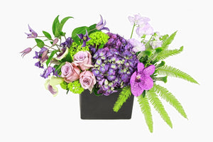 Flower arrangement in a low, square, black ceramic vase, purple hydrangea, purple clematis, purple vanda orchid, purple sweet peas and green viburnum