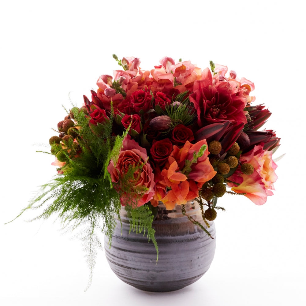 Flower arrangement in a low, round, brown, ceramic vase, coral roses, red spray roses, coral snapdragons, safari sunset and asparagus fern