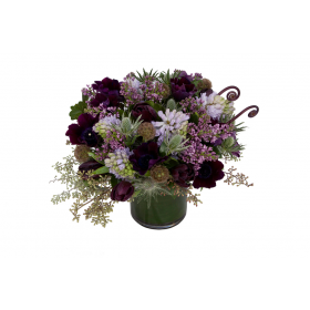 Flower arrangement in a low, round, clear glass vase, lilac, lavender hyacinths, purple anemones, purple tulips, scabiosa pods and seeded eucalyptus