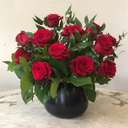 Flower arrangement in a low, round, black, ceramic vase, dozen red roses with lush greenery